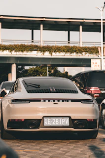 A luxury sports car is parked on a paved surface, positioned in front of a modern structure with greenery along the top. It is surrounded by other vehicles, indicating an urban setting. The car is sleek and shiny, with a focus on its rear design and a visible license plate.