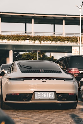 A luxury sports car is parked on a paved surface, positioned in front of a modern structure with greenery along the top. It is surrounded by other vehicles, indicating an urban setting. The car is sleek and shiny, with a focus on its rear design and a visible license plate.