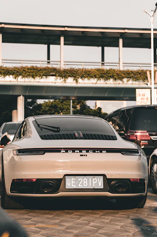 A luxury sports car is parked on a paved surface, positioned in front of a modern structure with greenery along the top. It is surrounded by other vehicles, indicating an urban setting. The car is sleek and shiny, with a focus on its rear design and a visible license plate.