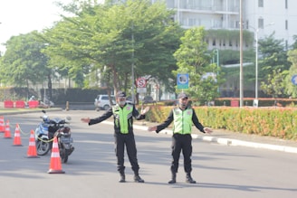 Two uniformed traffic officers stand on a road with traffic cones and a parked motorcycle nearby. They are directing traffic, with arms outstretched. The scene includes greenery and a building in the background on a sunny day.