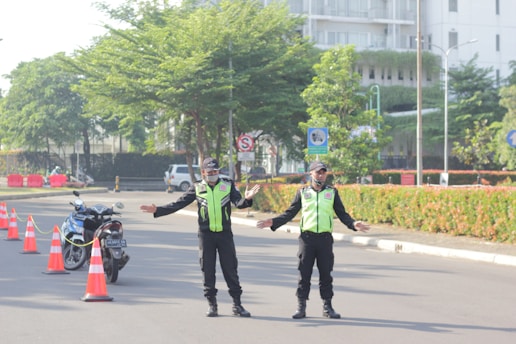 Two uniformed traffic officers stand on a road with traffic cones and a parked motorcycle nearby. They are directing traffic, with arms outstretched. The scene includes greenery and a building in the background on a sunny day.