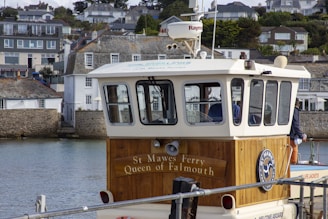 A ferry named St. Mawes Ferry Queen of Falmouth is docked near a coastal town. The ferry features a wooden section with the ferry's name inscribed on it. There are multiple residential buildings on the hillside in the background. The water is calm and reflects the buildings and ferry. The ferry has a navigation radar on top and windows around the cabin area.