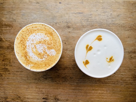 Two cups of coffee with intricate latte art designs on a wooden surface. One cup features sprinkled cinnamon on top, creating a speckled pattern over the foam, while the other showcases a delicate leaf pattern in the creamy foam.