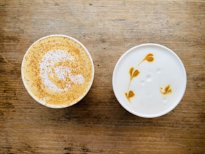 Two cups of coffee with intricate latte art designs on a wooden surface. One cup features sprinkled cinnamon on top, creating a speckled pattern over the foam, while the other showcases a delicate leaf pattern in the creamy foam.