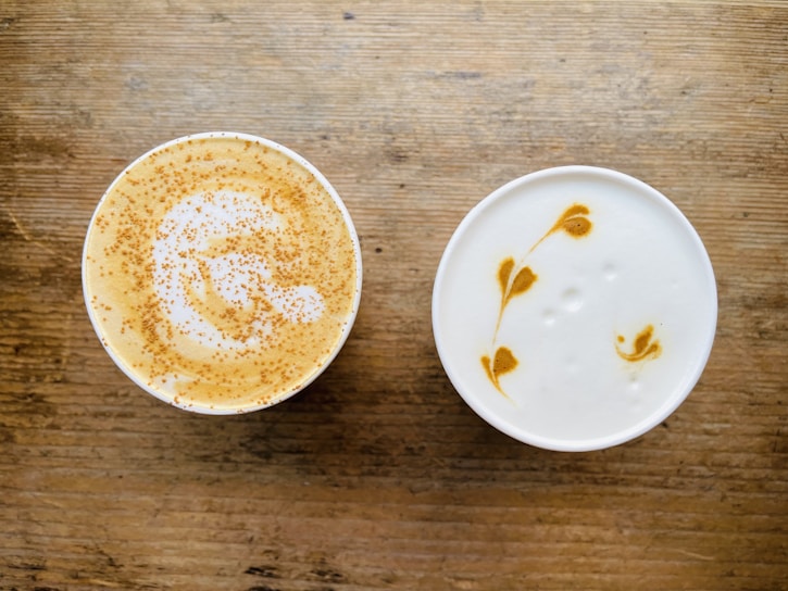 Two cups of coffee with intricate latte art designs on a wooden surface. One cup features sprinkled cinnamon on top, creating a speckled pattern over the foam, while the other showcases a delicate leaf pattern in the creamy foam.