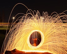 A mesmerizing display of light and movement created by long exposure photography. Bright, golden sparks form a circular pattern against a dark night sky. The sparks emanate from the center, creating arcs and trails of light. The scene conveys a sense of dynamic energy and creativity.