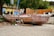A weathered and decaying old wooden boat rests on a gravel surface. The boat's paint is chipped and peeling, revealing patches of raw wood beneath. In the background, a wooden fence and colorful buildings can be seen alongside some greenery and trees.