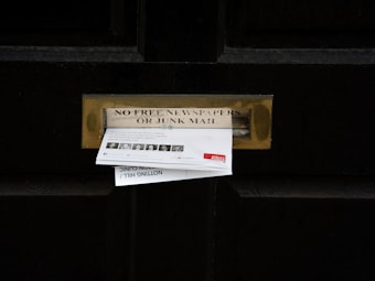 A black door with a brass mail slot containing two pieces of mail. Above the slot, a sign reads 'No Free Newspapers or Junk Mail'. The mail is protruding from the slot and includes text and images of people.