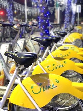 A row of yellow rental bicycles is neatly lined up, each featuring a smiley face logo and the word 'Ubike' on their rear wheel covers. The bicycles are parked on a street lined with trees wrapped in blue decorative lights, with more lights twinkling in the background. The scene is illuminated, suggesting it is nighttime.