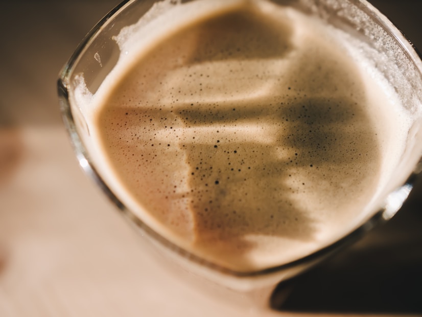 A close-up view of a glass filled with a creamy coffee drink, showcasing a thick layer of frothy foam on the top. The surface of the foam displays delicate speckles and variations in texture, illuminated by soft, warm lighting that casts subtle shadows.