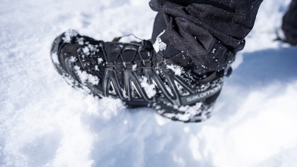 A close-up view of a black hiking boot covered in patches of snow, standing on a snowy surface. The snow is visibly packed around the boot, and the boot appears to be well-worn and suited for cold, snowy conditions.