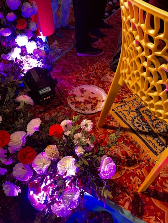 A vibrant and festive setting with bright artificial lighting illuminating an arrangement of red and white flowers. A decorative yellow chair is visible alongside a richly patterned red and gold carpet. There are scattered rose petals on the floor and a small plate also containing petals. People in the background are wearing varied attire, including sandals, but their faces are not visible.
