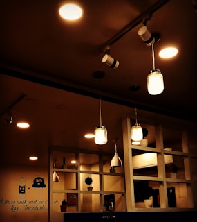 The image features a warmly lit indoor space with multiple light sources, including hanging pendant lights and ceiling lights. On the wall, there's a whimsical mural with a milk bottle and an ice cream character, alongside the text 'When milk met ice cream. Love. Inevitable.' A wooden shelving unit holds small potted plants and a few decorative items, contributing to the cozy and inviting atmosphere.