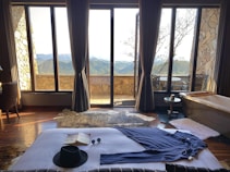 The image depicts a cozy room with large glass windows offering a scenic view of mountains. A bed with a black hat, an open book, and sunglasses is in the foreground. There is a stone wall, a bathtub to the right, and a table and chair set outside.