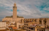 A large mosque with a tall minaret stands prominently above a cityscape with various residential buildings. The mosque features intricate architectural designs and is situated near the coast, visible in the background. The sky is partly cloudy with blue and gray tones.