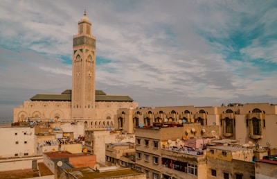 A large mosque with a tall minaret stands prominently above a cityscape with various residential buildings. The mosque features intricate architectural designs and is situated near the coast, visible in the background. The sky is partly cloudy with blue and gray tones.