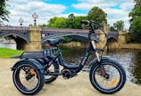 A tricycle bike with a black frame is parked on a concrete surface near a river. In the background, there is an ornate stone and metal bridge with elegant lampposts. The area is surrounded by lush green trees, and the sky above is partly cloudy.