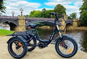 A tricycle bike with a black frame is parked on a concrete surface near a river. In the background, there is an ornate stone and metal bridge with elegant lampposts. The area is surrounded by lush green trees, and the sky above is partly cloudy.