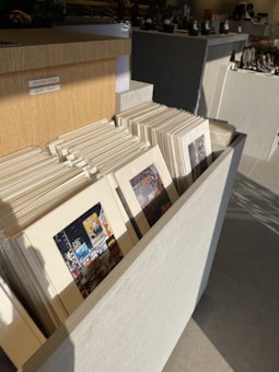 A well-lit room displays several framed pictures stacked vertically in a wooden holder. The pictures feature urban scenes, possibly recognizable landmarks or iconic cityscapes. A price tag indicates a promotion with a 'Buy 2 Get 1 Free' offer. Shadows cast on the floor suggest sunlight entering from a nearby window.