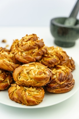 A plate of freshly baked pastries topped with sliced almonds is displayed. The pastries have a golden-brown crust and appear flaky and buttery. In the background, there is a mortar and pestle set with a stone texture.