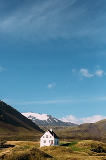 A solitary white house with a dark roof sits in a lush, green landscape surrounded by grass-covered hills. In the background, there are snow-capped mountains under a bright blue sky with some scattered clouds.