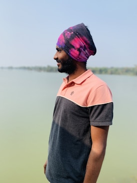 A man standing sideways near a serene body of water, wearing a colorful turban and a polo shirt. The background is a calm, expansive lake with a clear sky above.