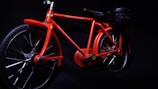 A vibrant red toy bicycle is displayed against a dark background. It features a detailed frame, with visible text on the top bar indicating 'MOVEMENT MAGIC POWER' and 'ELECTRICITY MOVES THE BICYCLE'. The bicycle includes two large wheels and a small attachment at the rear resembling a battery or motor pack.