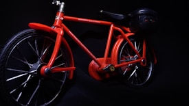 A vibrant red toy bicycle is displayed against a dark background. It features a detailed frame, with visible text on the top bar indicating 'MOVEMENT MAGIC POWER' and 'ELECTRICITY MOVES THE BICYCLE'. The bicycle includes two large wheels and a small attachment at the rear resembling a battery or motor pack.