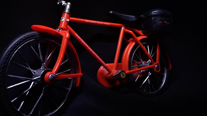 A vibrant red toy bicycle is displayed against a dark background. It features a detailed frame, with visible text on the top bar indicating 'MOVEMENT MAGIC POWER' and 'ELECTRICITY MOVES THE BICYCLE'. The bicycle includes two large wheels and a small attachment at the rear resembling a battery or motor pack.
