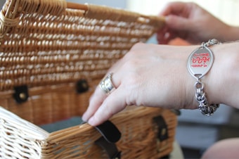 A close-up view of a person's hands handling a wicker basket. The individual is wearing a silver bracelet with red engraved text and a silver ring on one of the fingers. The wicker basket features leather straps and metal buckles.