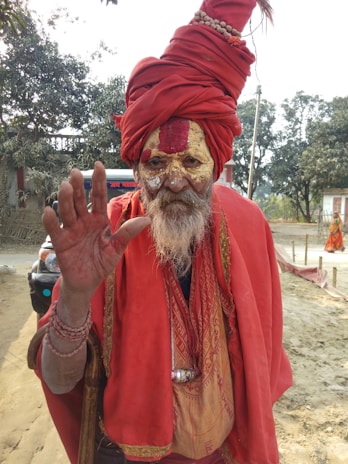 A person dressed in traditional attire with a red turban and robe. The face is painted with intricate designs, and they are raising a hand, showing bracelets. The background includes trees and a vehicle.