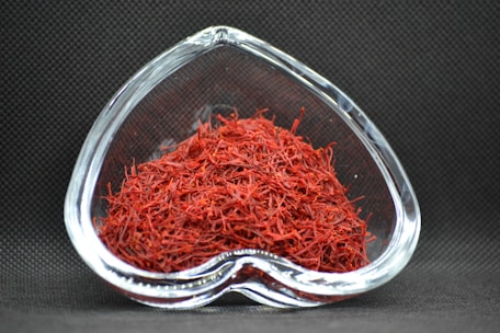 A heart-shaped clear glass bowl filled with red saffron threads, set against a dark textured background. The vibrant red color of the saffron contrasts with the dark setting, highlighting the intricate strands.