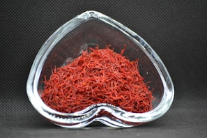 A heart-shaped clear glass bowl filled with red saffron threads, set against a dark textured background. The vibrant red color of the saffron contrasts with the dark setting, highlighting the intricate strands.