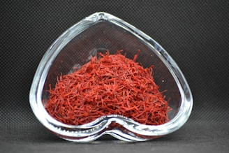 A heart-shaped clear glass bowl filled with red saffron threads, set against a dark textured background. The vibrant red color of the saffron contrasts with the dark setting, highlighting the intricate strands.