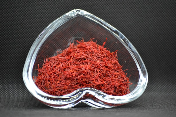A heart-shaped clear glass bowl filled with red saffron threads, set against a dark textured background. The vibrant red color of the saffron contrasts with the dark setting, highlighting the intricate strands.