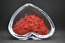 A heart-shaped clear glass bowl filled with red saffron threads, set against a dark textured background. The vibrant red color of the saffron contrasts with the dark setting, highlighting the intricate strands.