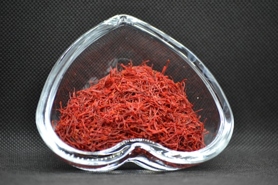 A heart-shaped clear glass bowl filled with red saffron threads, set against a dark textured background. The vibrant red color of the saffron contrasts with the dark setting, highlighting the intricate strands.