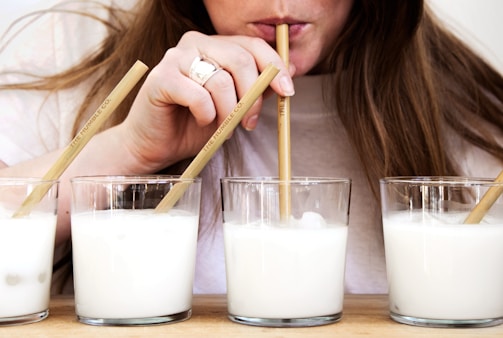 A person is sipping from a glass filled with a creamy white liquid using a bamboo straw. Three additional glasses filled with the same liquid are placed on a wooden surface. The person has long hair and is wearing a white shirt. The scene gives a casual and relaxed impression.