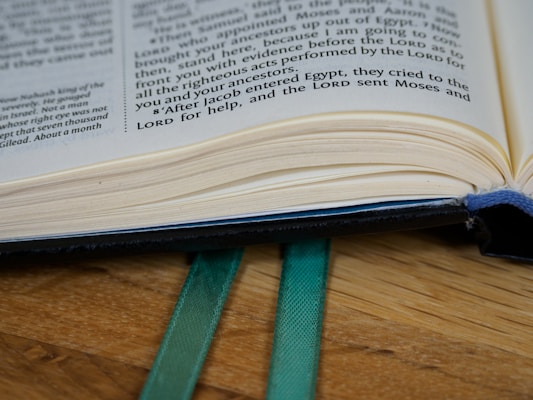An open book resting on a wooden surface with visible text on the pages and two green ribbons protruding from the bottom.