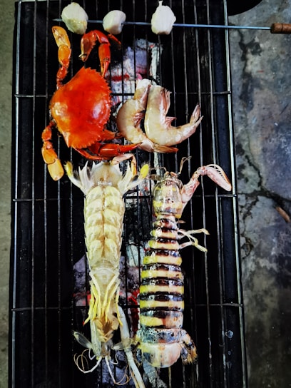 A variety of seafood including a red crab, two large prawns, and a lobster are placed on a grill over smoldering coals. Above them, skewered pieces of food, possibly scallops or mushrooms, are also being grilled. The grilling surface is black with visible grill marks and the coals below emit a reddish glow.