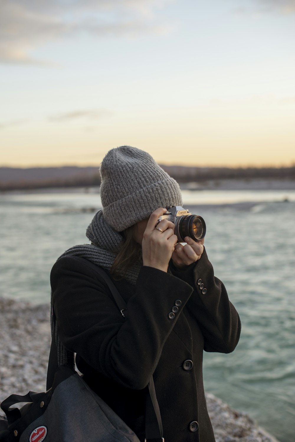 person in gray knit cap and black jacket taking photo of body of water during daytime