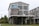 A two-story house on stilts with large windows and a blue metal roof. The upper level features a wraparound deck with chairs, and there is an open area beneath the house. The structure is surrounded by grassy landscaping and neighboring houses.
