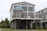 A two-story house on stilts with large windows and a blue metal roof. The upper level features a wraparound deck with chairs, and there is an open area beneath the house. The structure is surrounded by grassy landscaping and neighboring houses.