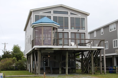 A two-story house on stilts with large windows and a blue metal roof. The upper level features a wraparound deck with chairs, and there is an open area beneath the house. The structure is surrounded by grassy landscaping and neighboring houses.