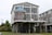 A two-story house on stilts with large windows and a blue metal roof. The upper level features a wraparound deck with chairs, and there is an open area beneath the house. The structure is surrounded by grassy landscaping and neighboring houses.