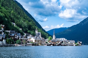 A picturesque lakeside village is nestled between steep, forested mountains. The village consists of traditional European buildings, many with a distinct alpine architectural style. A prominent church with a tall, pointed spire is centrally located, enhancing the quaint charm. The water reflects the skyline, and the sky is partly cloudy with a deep blue hue.