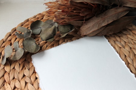 A natural and rustic scene featuring a woven placemat as the base. On top of it lies a blank sheet of paper with a rough edge, surrounded by dried eucalyptus leaves and a dried protea flower. The colors are muted, giving the composition a calm and organic feel.