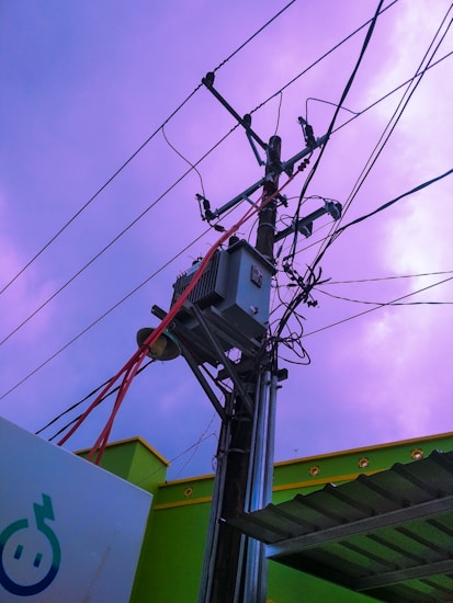 A utility pole with numerous electrical wires is prominently featured, set against a sky with shades of purple. An electrical transformer is mounted on the pole, with red and black wires extending outward. Buildings with vibrant green walls and an ornamental rooftop edge are visible in the background.