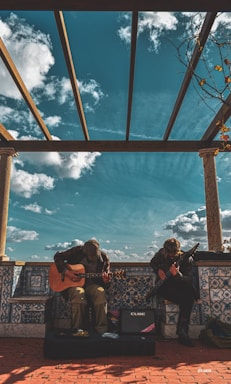 Two individuals are seated, playing guitars under a pergola with a vivid blue sky filled with scattered clouds. The scene is set on a tiled platform with decorative patterns, and they are accompanied by a small amplifier. Bare branches with sparse leaves frame the top right corner.