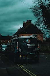A person stands at the back of a dark-colored service van on a cloudy day. The background includes residential buildings and bare tree branches against an overcast sky. The scene appears to take place on a street with additional vehicles parked along the side.